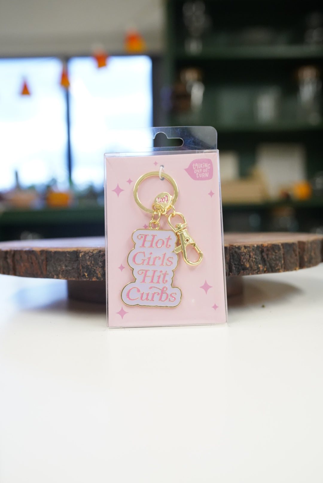 A gold-tone metal keychain with a pink enamel sign that reads 'Hot Girls Hit Curbs' in white lettering, presented in its original pink packaging card on a white tabletop with a blurred background.