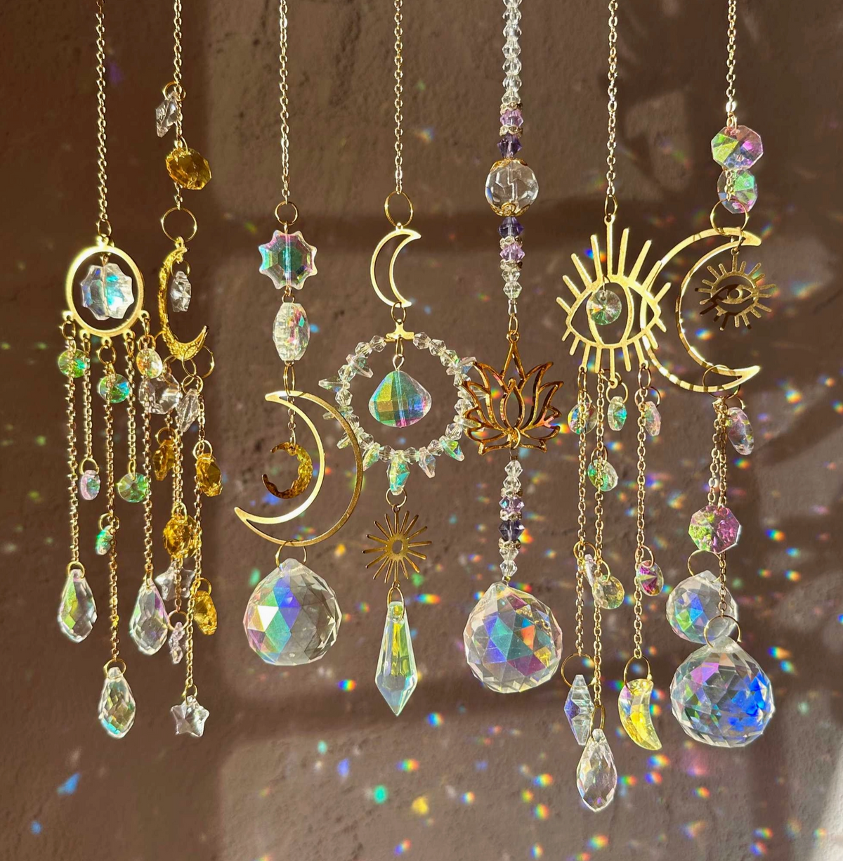 A set of gold-toned hanging decorative ornaments with crescent moons, suns, eye motifs, and faceted iridescent crystal drops, arranged against a warm brown background with rainbow light reflections.