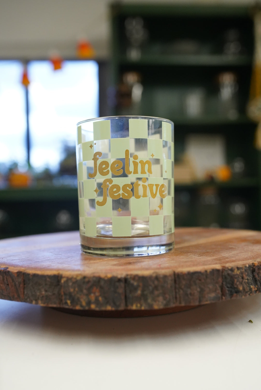 A clear glass tumbler with a pale green checkerboard pattern and gold foil lettering that reads 'feelin' festive', displayed on a rustic wood slice coaster with a blurred indoor background.